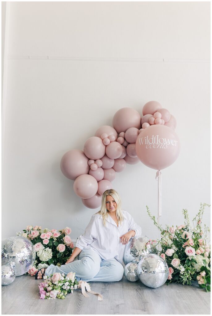 Morgan sitting on the floor surrounded by floral arrangements and a balloon arch, celebrating the anniversary of Wildflower Events in a relaxed, creative setup.