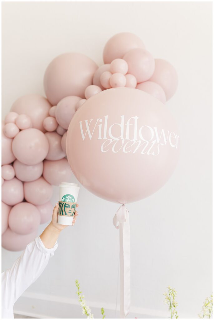 Close-up of a custom balloon with 'Wildflower Events' printed on it, part of a celebratory balloon arch setup in soft, neutral tones.