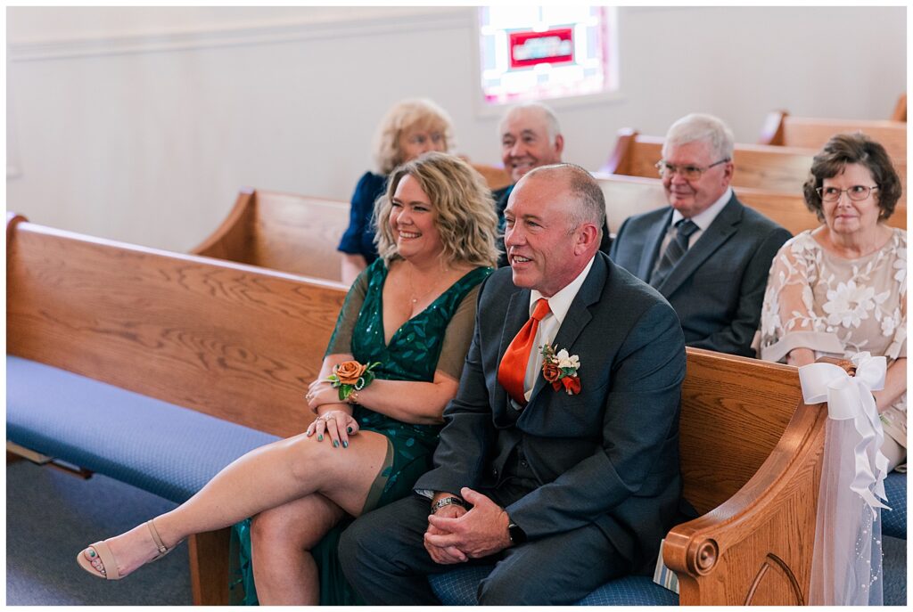Guests smiling and watching the ceremony from the front row of the church, with corsages and boutonnières highlighting their roles.