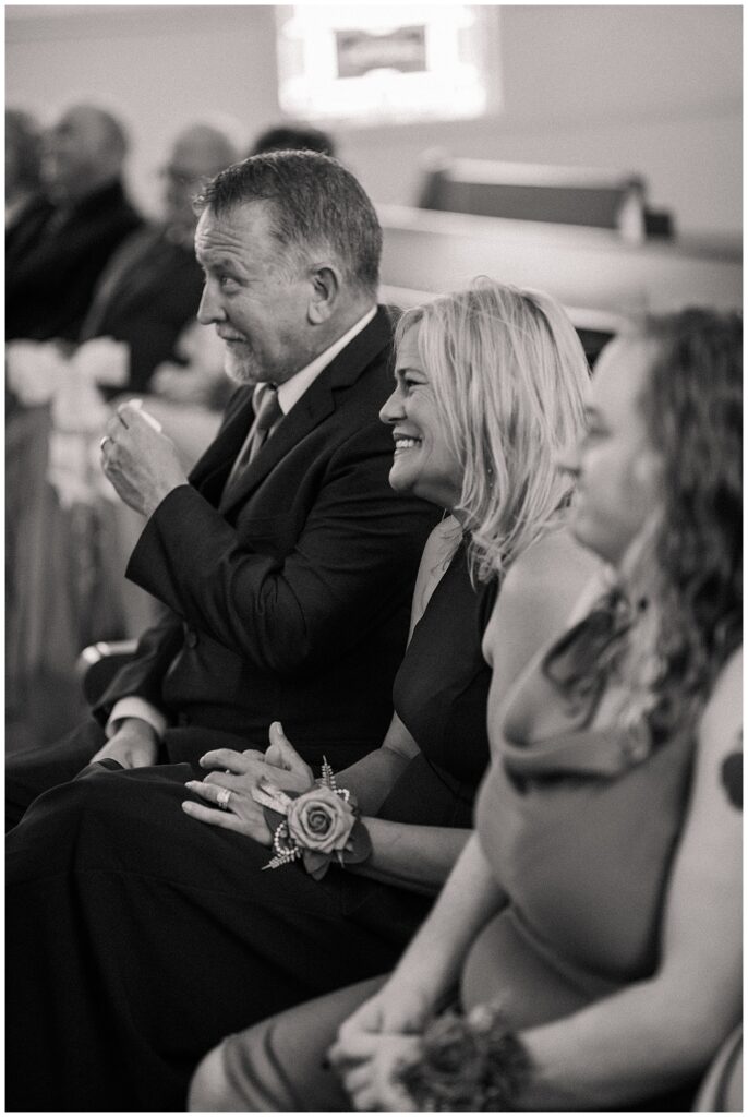 Black and white image of guests laughing and reacting during the wedding ceremony from the pews.