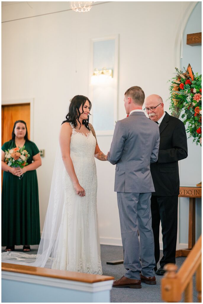 Bride and groom exchanging rings during their wedding ceremony, with a bridesmaid and officiant looking on.