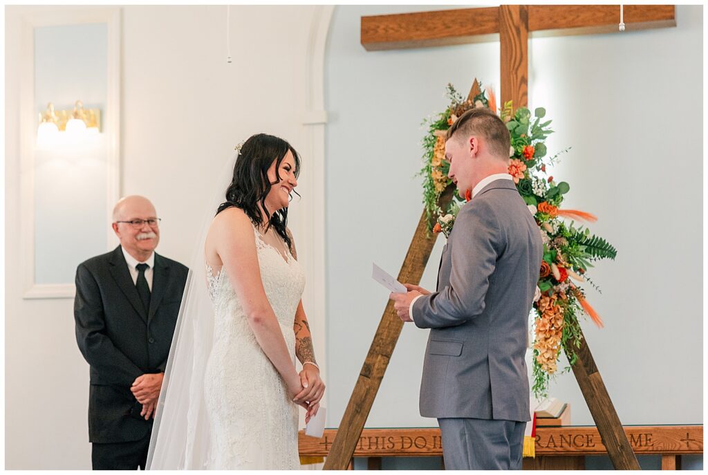 Bride smiling as she listens to the groom read his vows during their wedding ceremony at the altar.