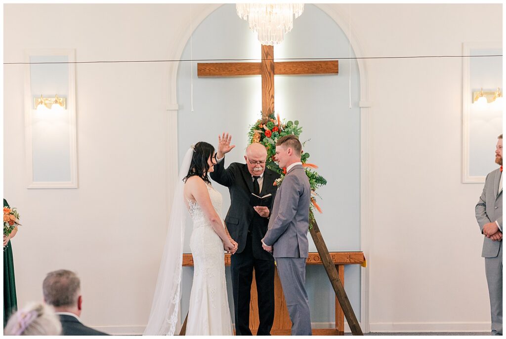 Bride and groom holding hands at the altar as the officiant prays over them during their church wedding ceremony.