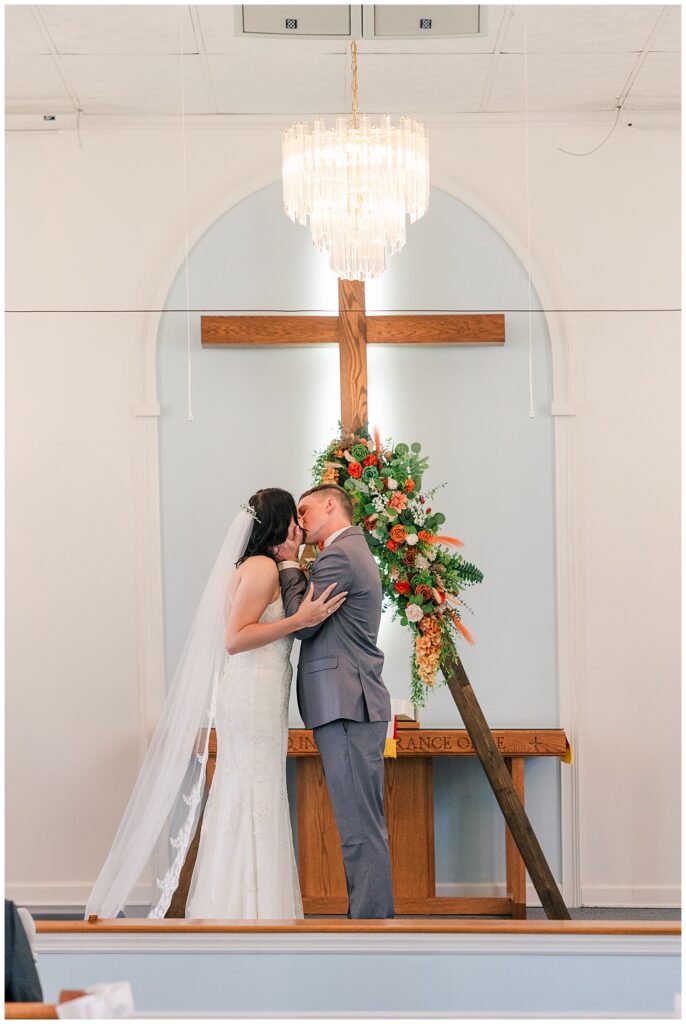 Bride and groom sharing their first kiss in front of a large wooden cross decorated with florals at the altar of their church ceremony.