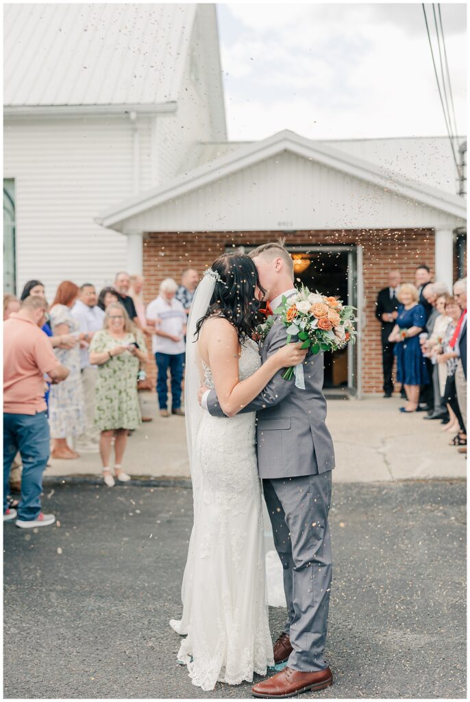 Bride and groom kissing outside a small white church after the ceremony, surrounded by cheering family and friends.