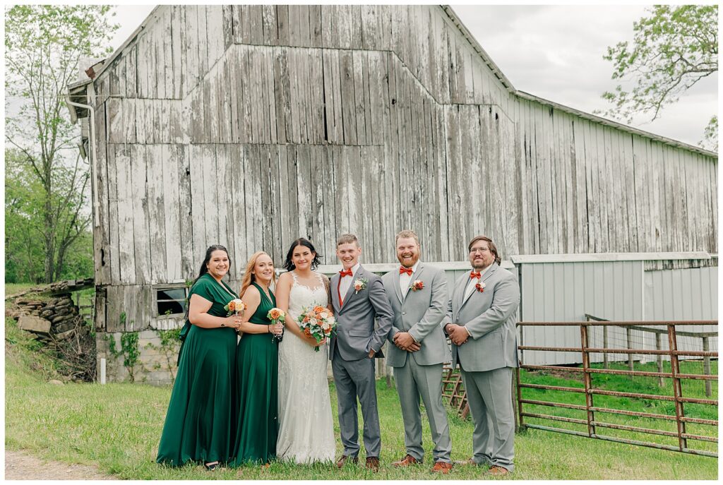 Bride and groom posing with their wedding party in front of a rustic white barn, all smiling on a grassy lawn.