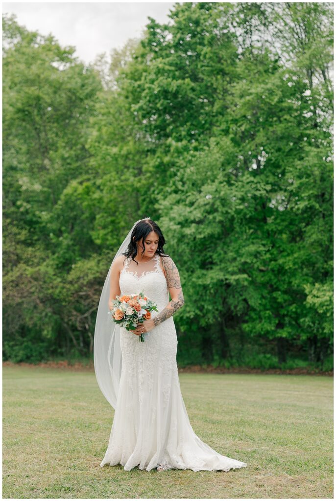 Bride standing alone in a grassy field, holding her bouquet and smiling softly in front of lush greenery.