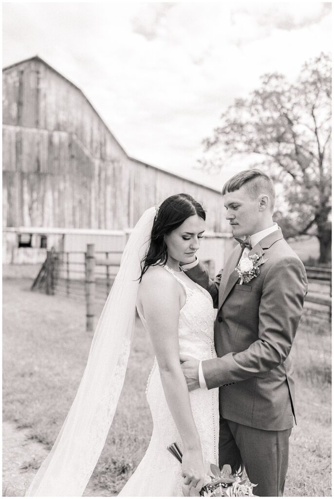 Black and white photo of the bride resting her head on the groom’s chest as they embrace in front of a rustic barn.