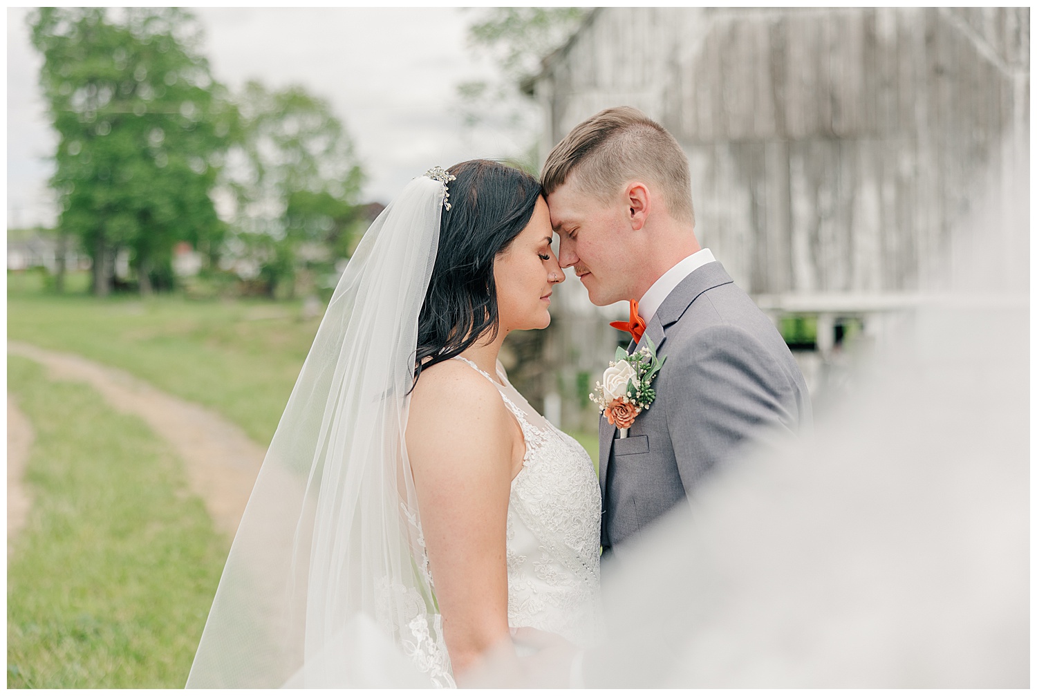 Bride and groom standing forehead to forehead in front of a rustic barn on a country road, captured in a soft, romantic moment on their wedding day.