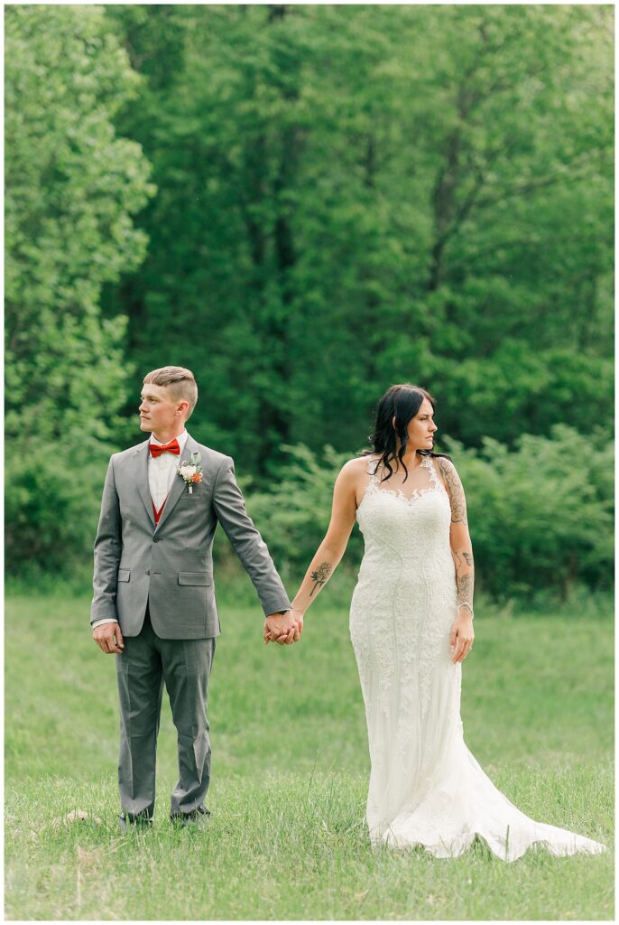 Bride and groom holding hands while walking through a grassy field surrounded by trees, captured in a candid moment.