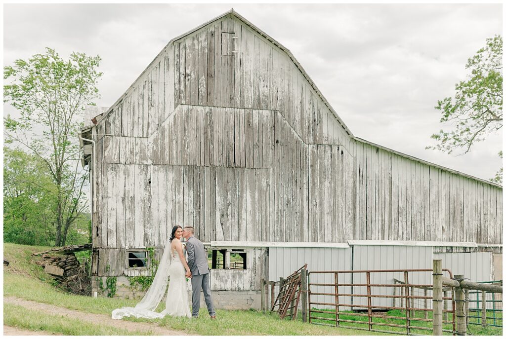 Bride and groom holding hands in front of a weathered white barn, posing together on a lush green lawn.