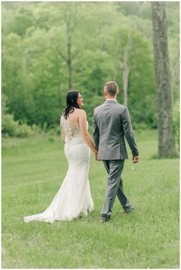 Bride and groom holding hands while walking through a grassy field, captured from behind in a quiet, romantic moment.