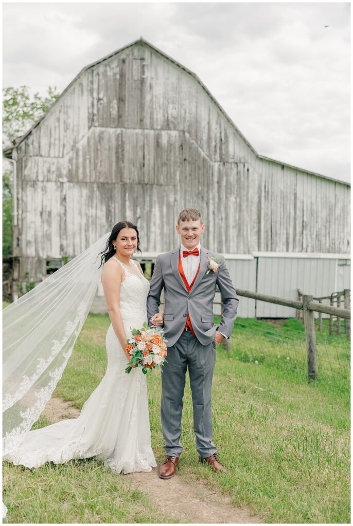 Bride and groom posing with smiles in front of a rustic wooden barn, holding a colorful bouquet of flowers.