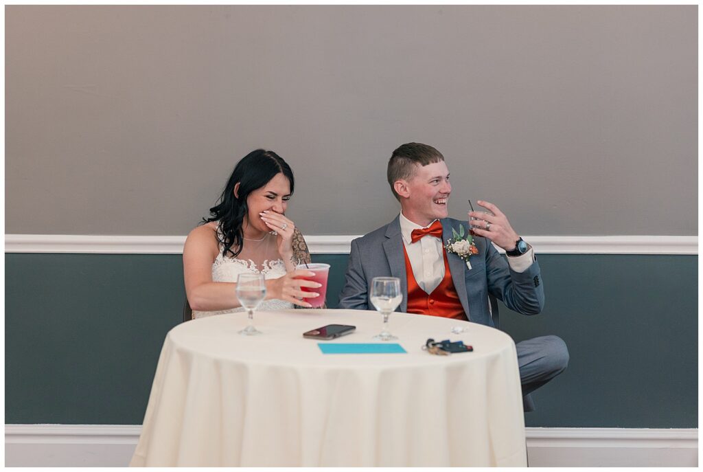 Bride and groom laughing and enjoying drinks together while seated at their sweetheart table during the reception.