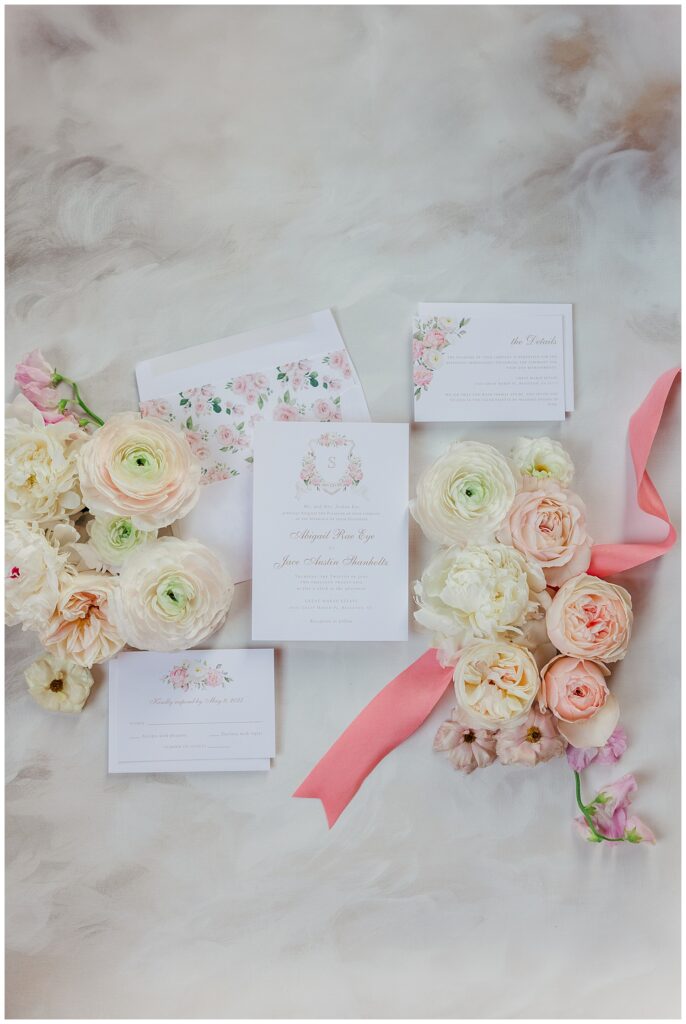 Styled wedding invitation suite surrounded by pastel florals and pink ribbon, showcasing the soft pink color palette of the day.