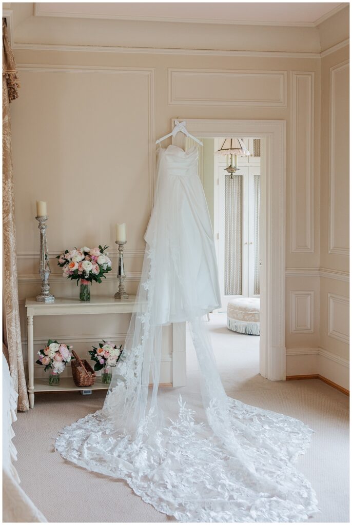 Bridal gown hanging elegantly in the bridal suite next to a floral display and antique mirror at Great Marsh Estate.