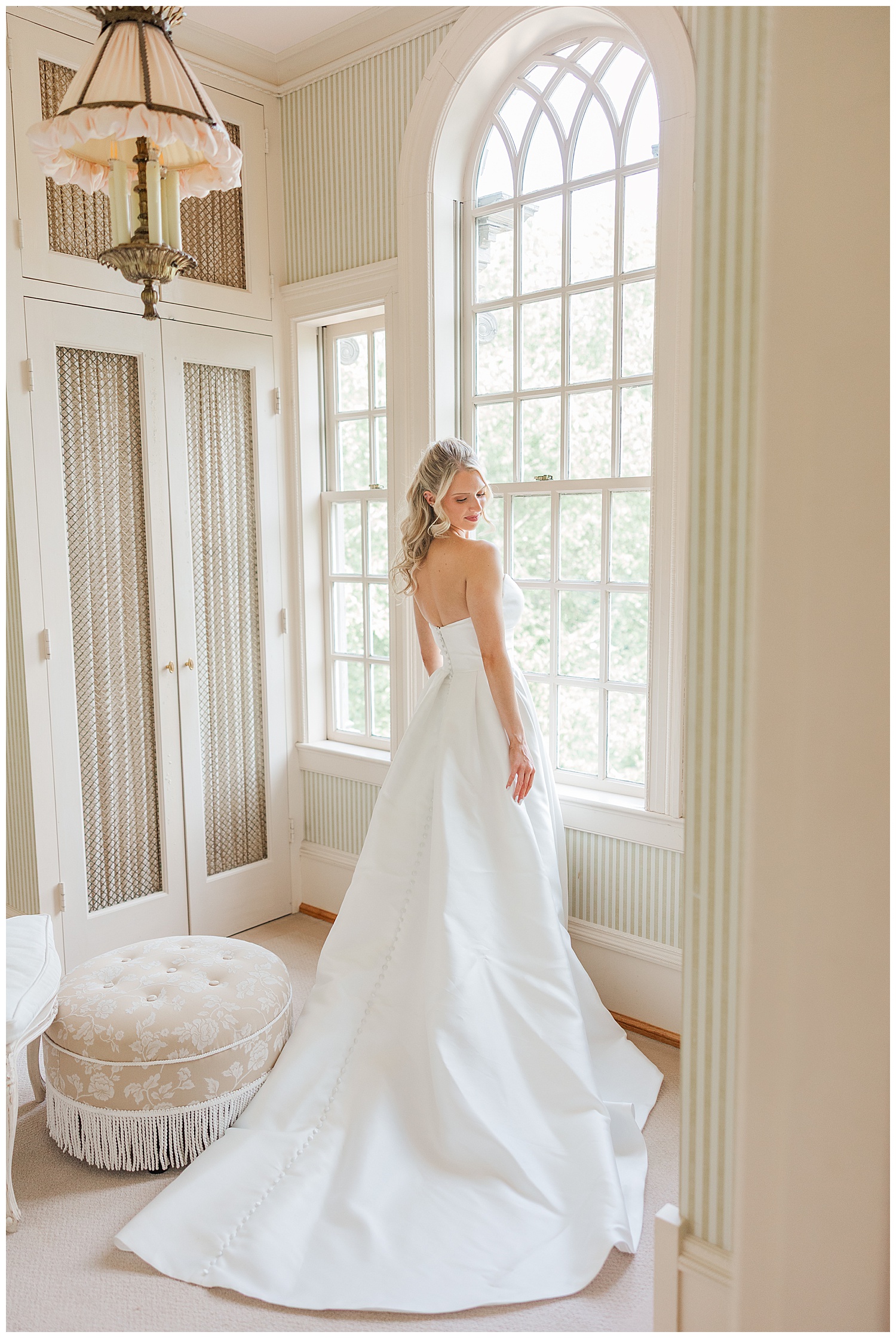 Bride stands in front of tall windows, holding her gown and smiling before the ceremony at Great Marsh Estate.