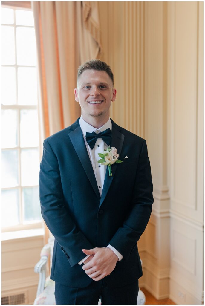 Groom poses by a bright window in a navy tuxedo, smiling just before the ceremony begins.
