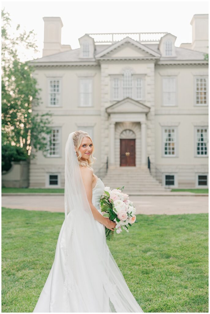 Bride smiles brightly while holding her bouquet with her full train fanned out, framed by the classic columns of Great Marsh Estate.
