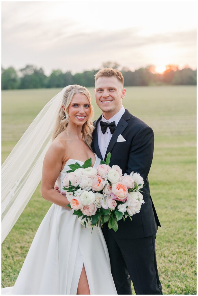Bride and groom smile at sunset in an open field, the bride holding a lush bouquet of blush and ivory florals with her veil cascading behind her. Zoe Evans Photography.