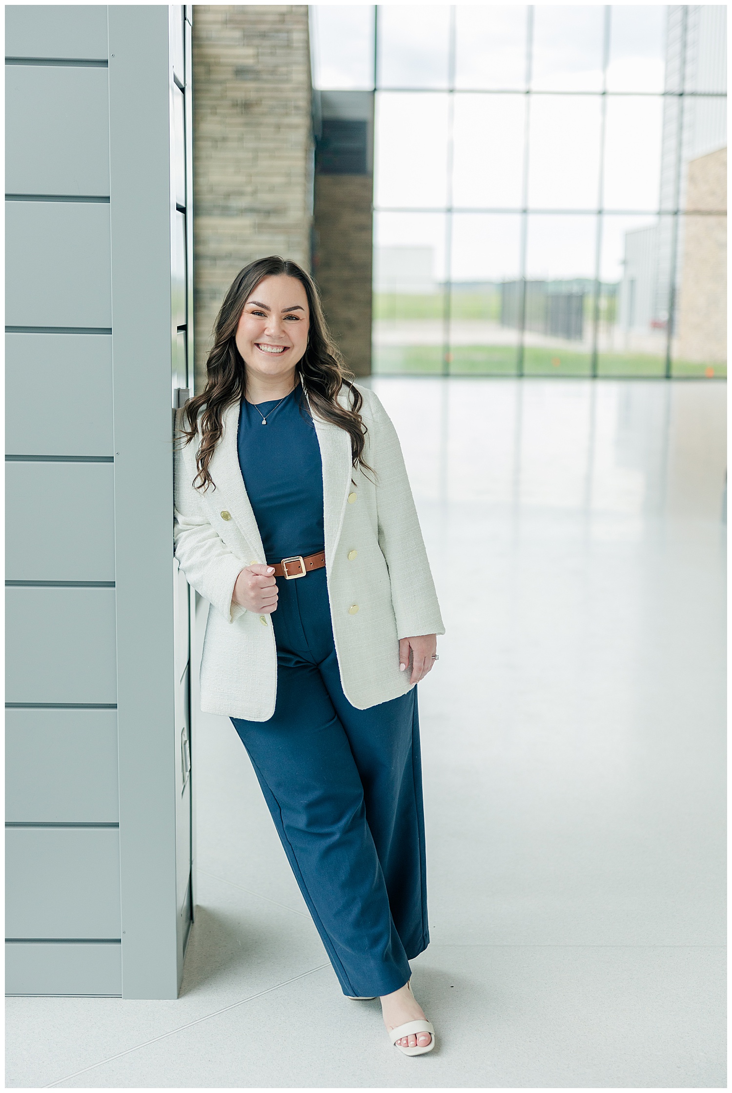 Professional woman in a white blazer and navy jumpsuit standing confidently by modern glass doors.