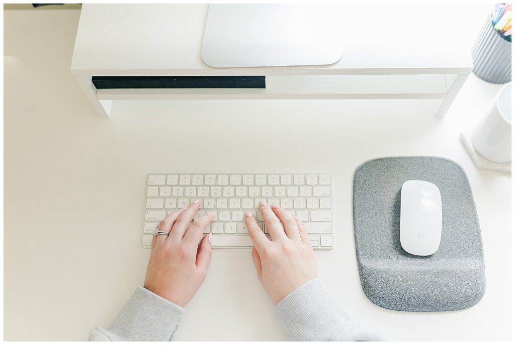 Overhead view of woman’s hands typing on a white keyboard at a minimal, clean desk setup.
