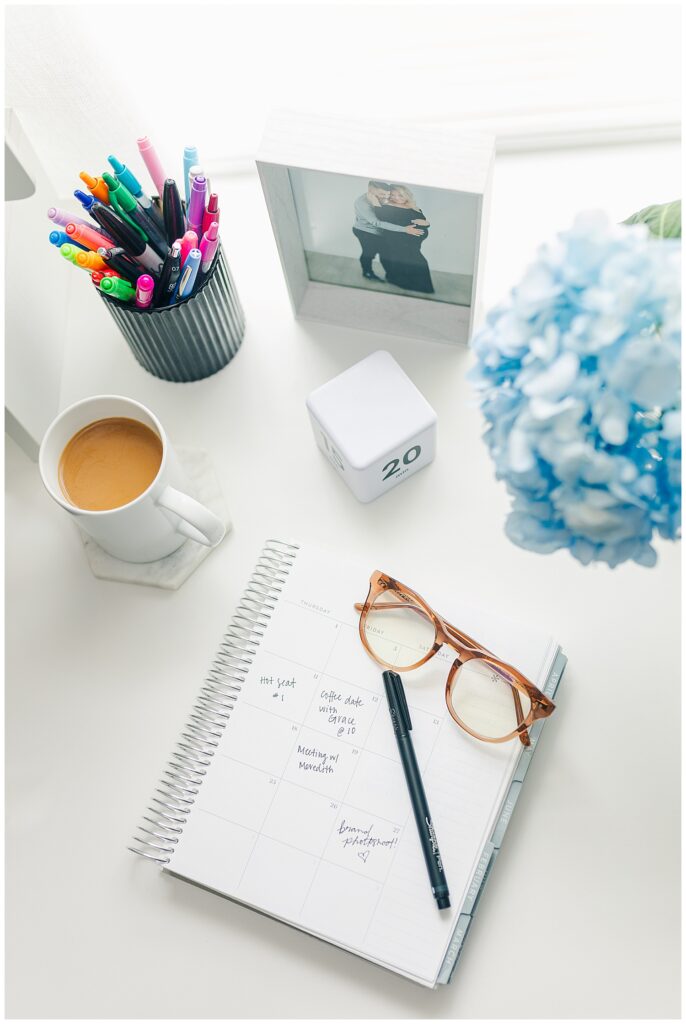 Flat lay with coffee, glasses, pens, planner, and blue flowers on a creative workspace.