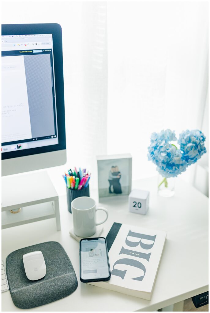 Modern workspace setup with computer, coffee, flowers, and office accessories on a bright white desk. Zoe Evans Photography.