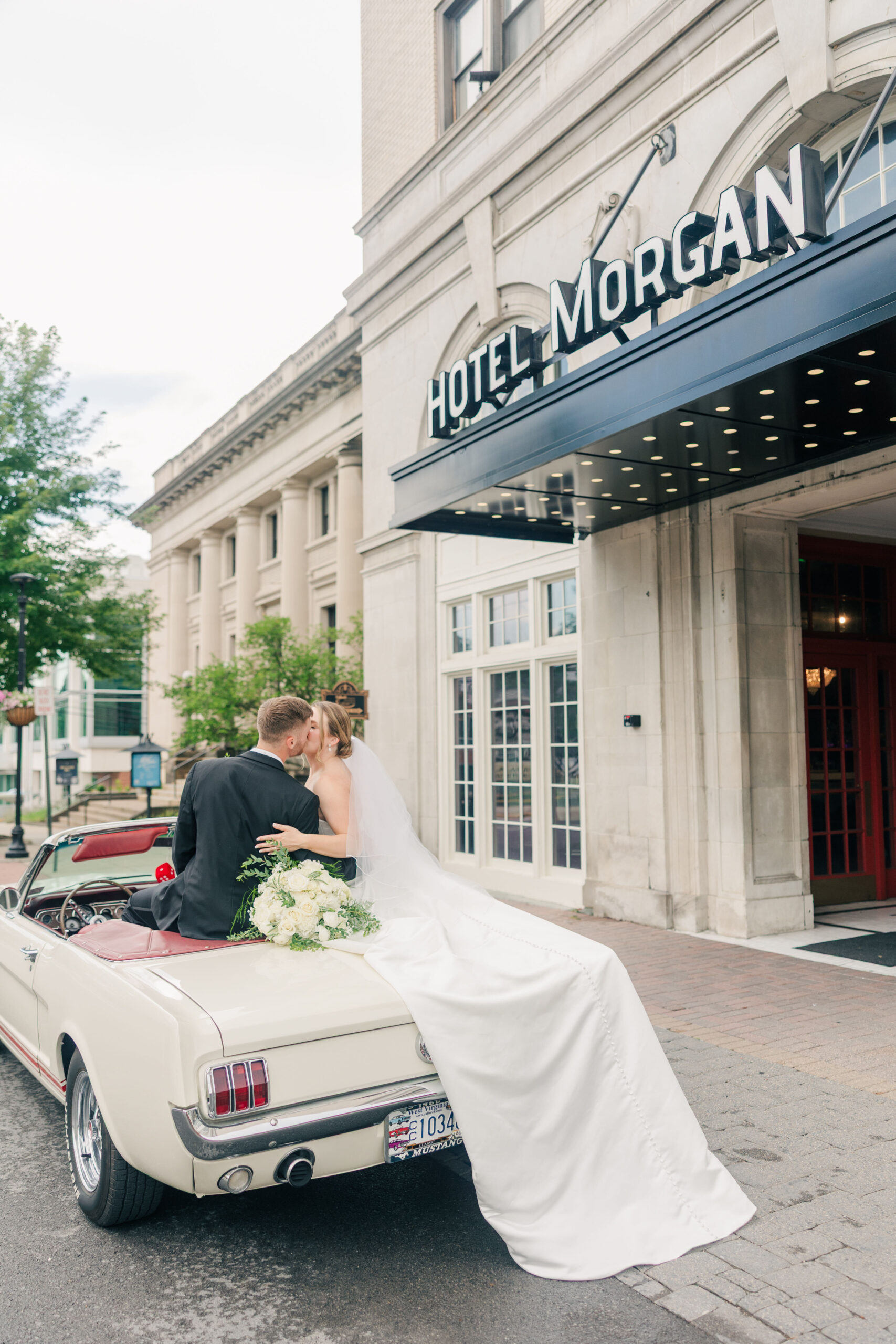 Bride and groom sit on the back of a vintage white Mustang outside Hotel Morgan in Morgantown, West Virginia, sharing a quiet moment after their wedding ceremony.
