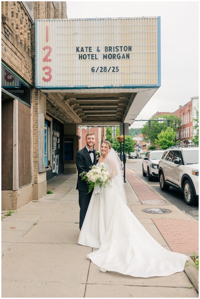 Bride and groom smile outside a vintage movie theater marquee that reads “Hotel Morgan – 6/28/25” on the sidewalk in Morgantown.