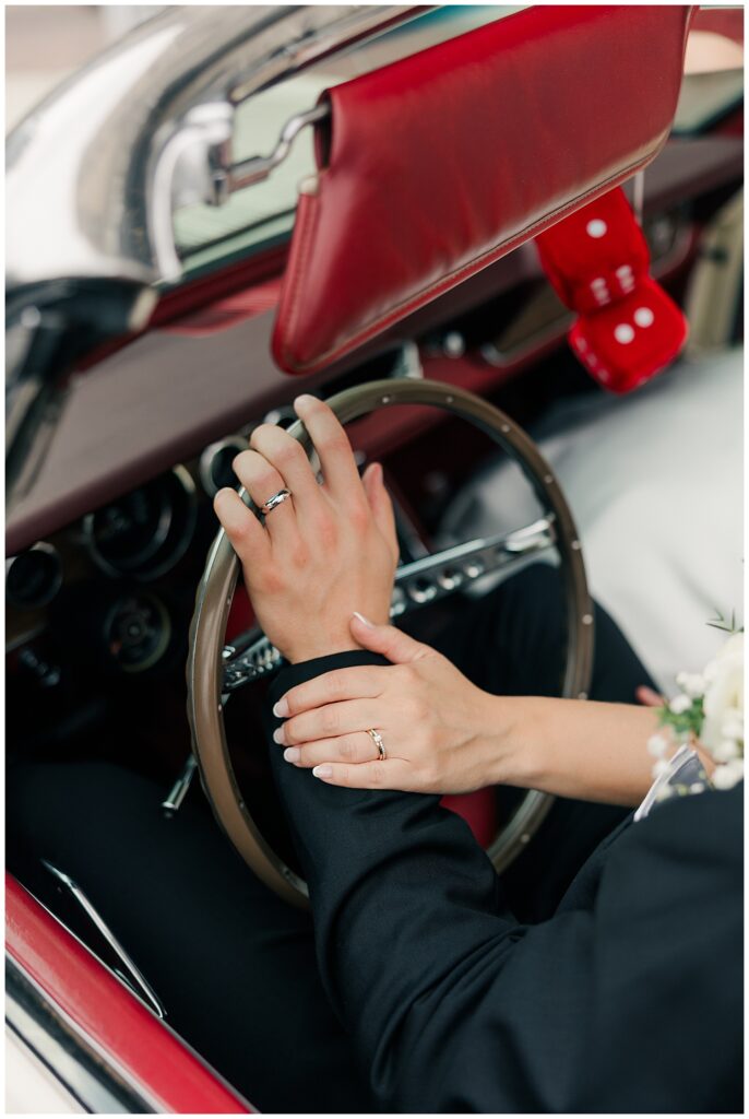 Close-up of the bride and groom’s hands on the steering wheel of their vintage getaway car, showing off wedding rings and bouquet detail.