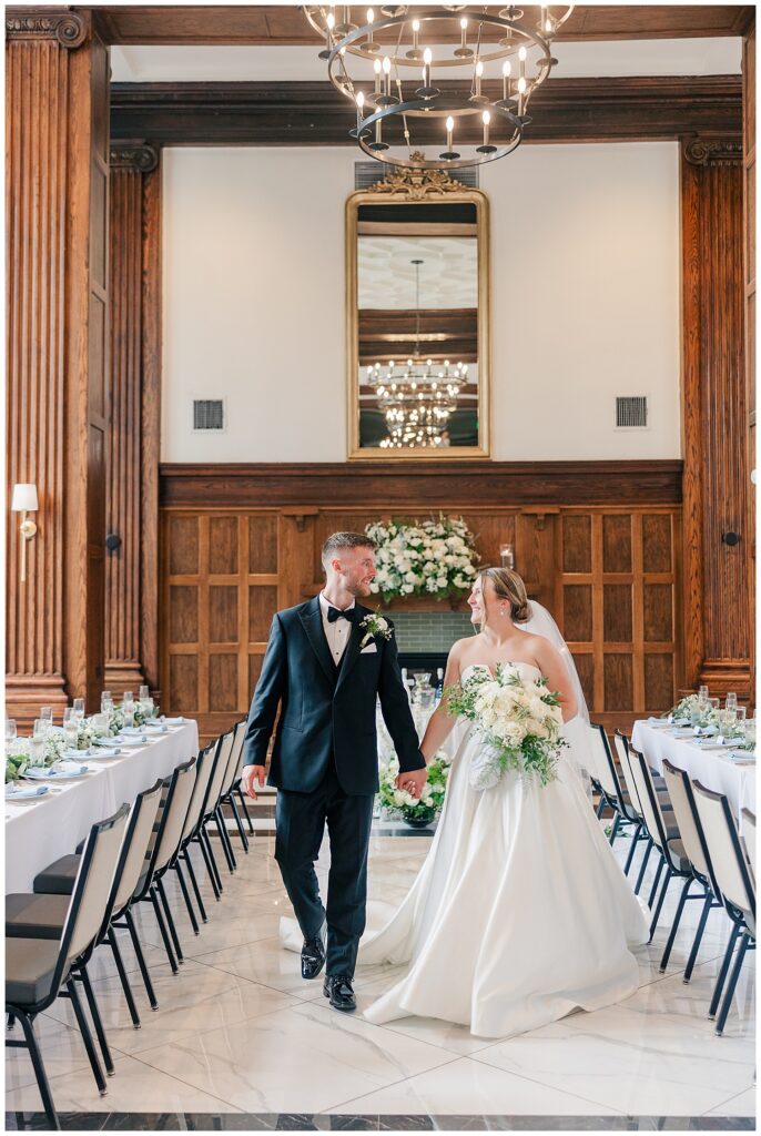 Bride and groom walk hand-in-hand through their elegant reception space at Hotel Morgan, surrounded by long tables with white linens and greenery.
