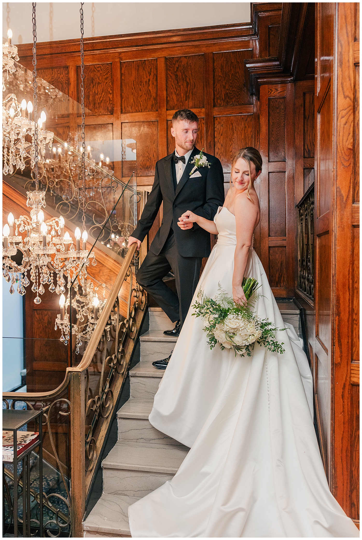Bride and groom pose on a grand chandelier-lit staircase inside Hotel Morgan, the bride holding a classic white bouquet.