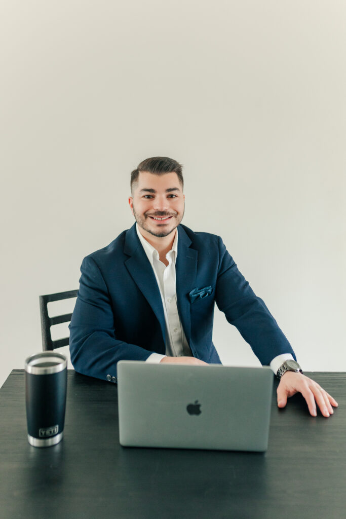 Male entrepreneur in a navy blazer working on a laptop at a black desk with a tumbler nearby