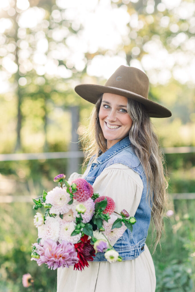 Woman in a wide-brim hat and denim vest smiling while holding a bouquet of fresh flowers outdoors