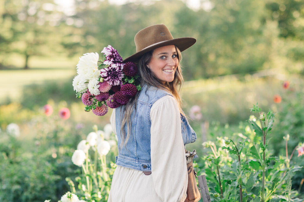 Woman wearing a hat and denim vest walking through a flower field with a colorful bouquet over her shoulder
