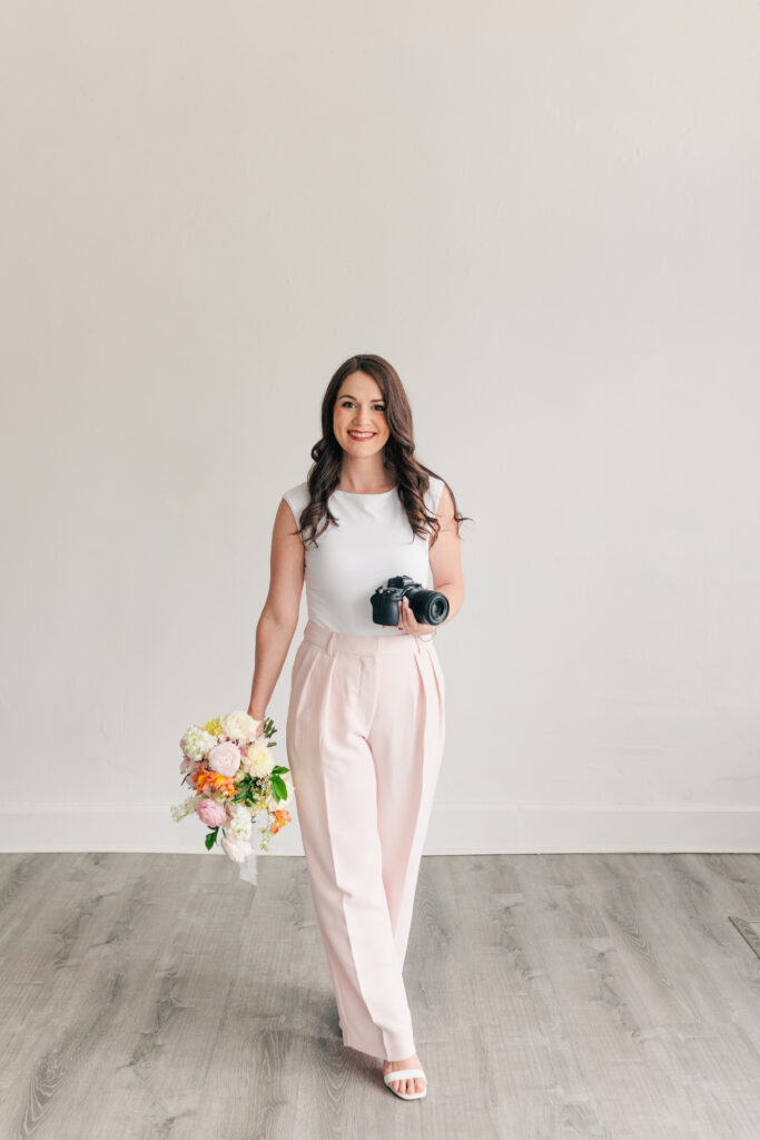 Female creative entrepreneur holding flowers and a camera, walking in a bright studio space