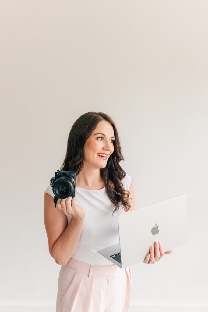 Female brand photographer smiling while holding a camera and working on a laptop