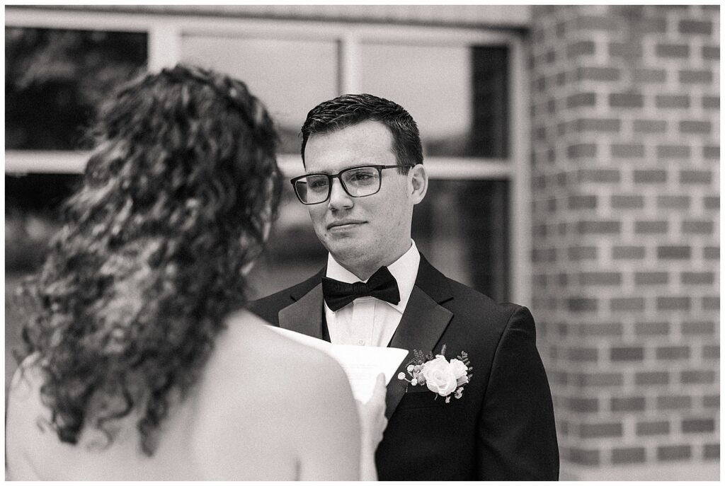 Black and white close-up of groom looking at bride while she reads her vows