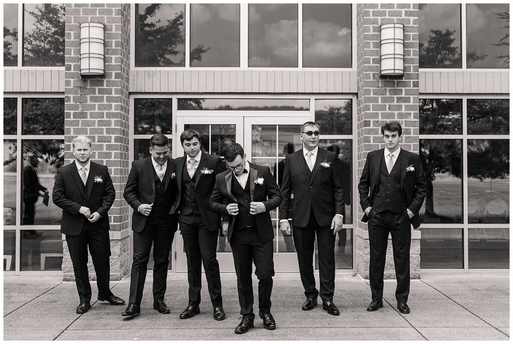 Black and white photo of groom and groomsmen laughing in front of modern brick venue
