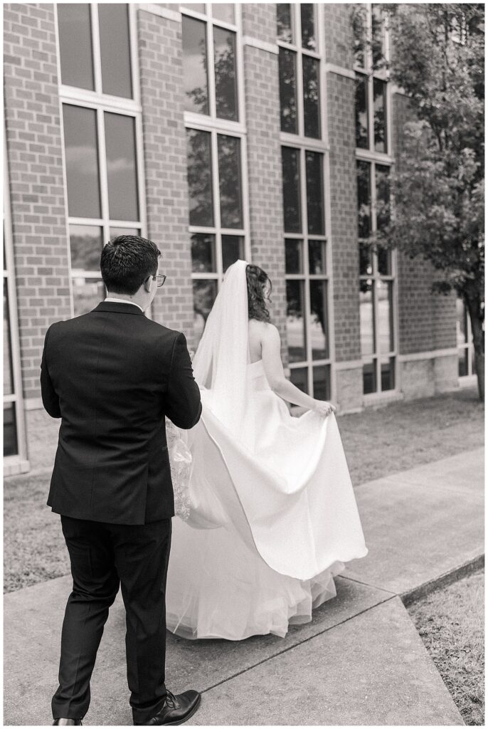 Black and white image of groom watching bride walk in front of a brick building
