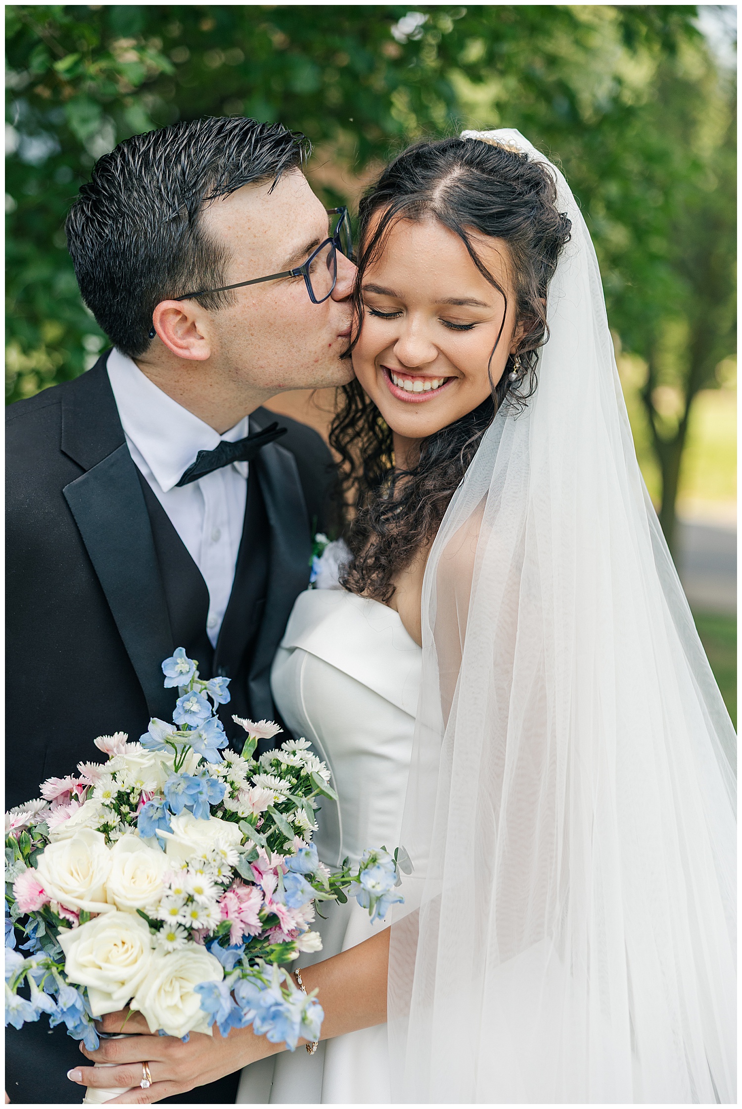 Bride and groom smile as he kisses her cheek, holding a soft pastel bouquet