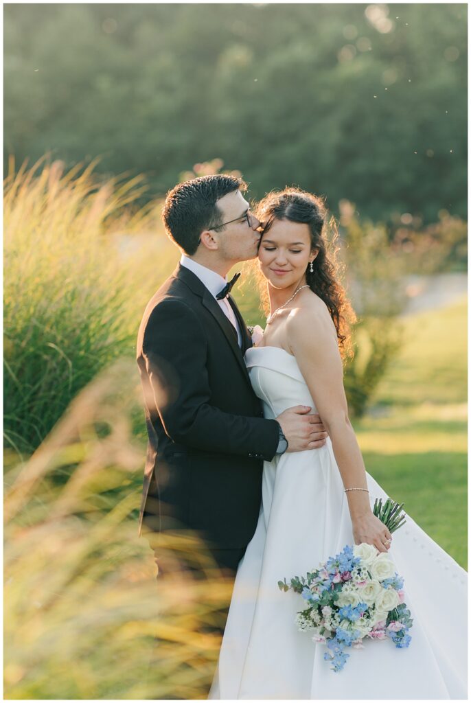 Groom kisses bride’s temple as they stand in a sunlit field of tall grass — captured by Zoe Evans Photography