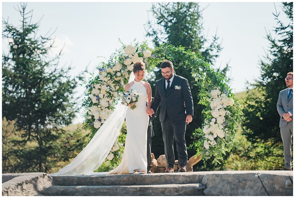 Bride and groom walk down stone steps after their outdoor ceremony at Red Barn Inn, surrounded by lush greenery and white floral arch