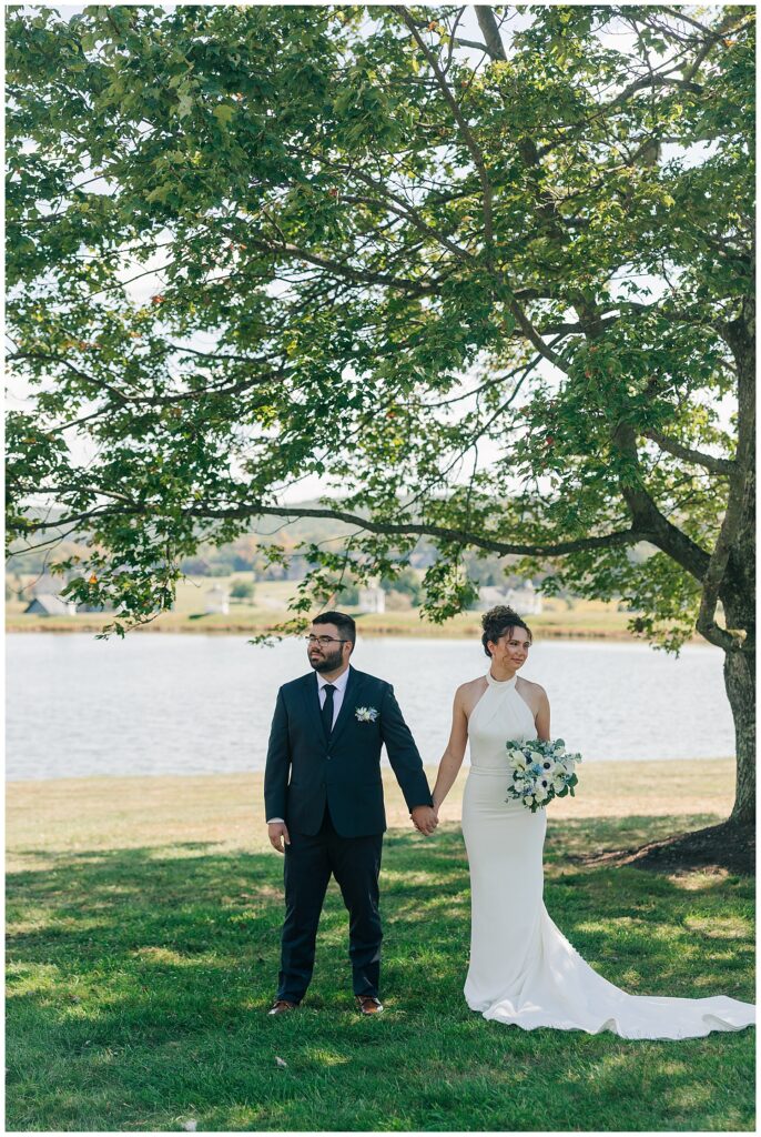 Bride and groom stand hand in hand beneath shady tree with lake in background at Deep Creek wedding venue