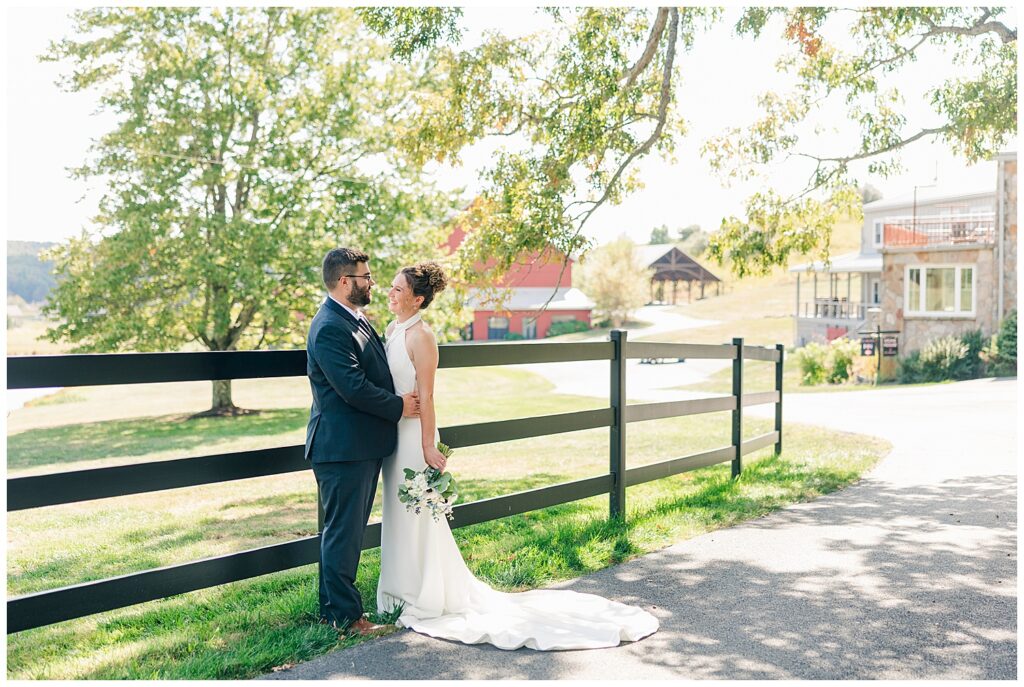 Bride and groom embrace along a rustic black fence line at Red Barn Inn with trees and barn in the distance