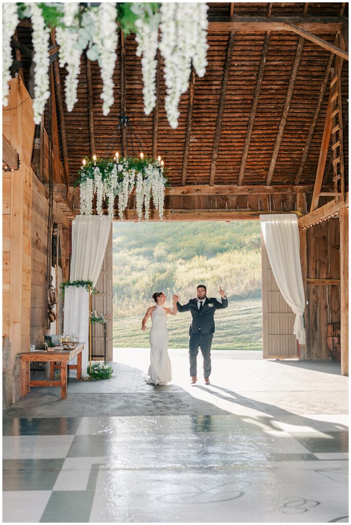 Bride and groom entering their reception with joyful energy, arms raised under rustic barn beams and floral ceiling installation