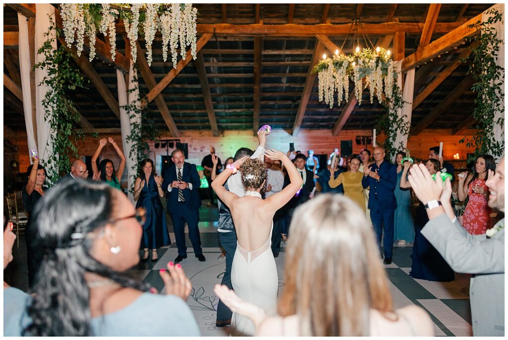 Bride with arms raised dancing at her wedding reception surrounded by cheering guests under floral chandeliers – Zoe Evans Wedding Photography