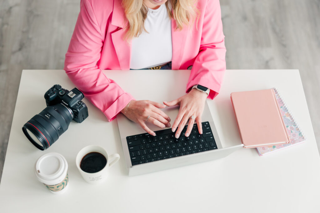 Woman in pink blazer typing on laptop with camera gear nearby – styled flat lay brand photos by Zoe Evans Photography for creative professionals.