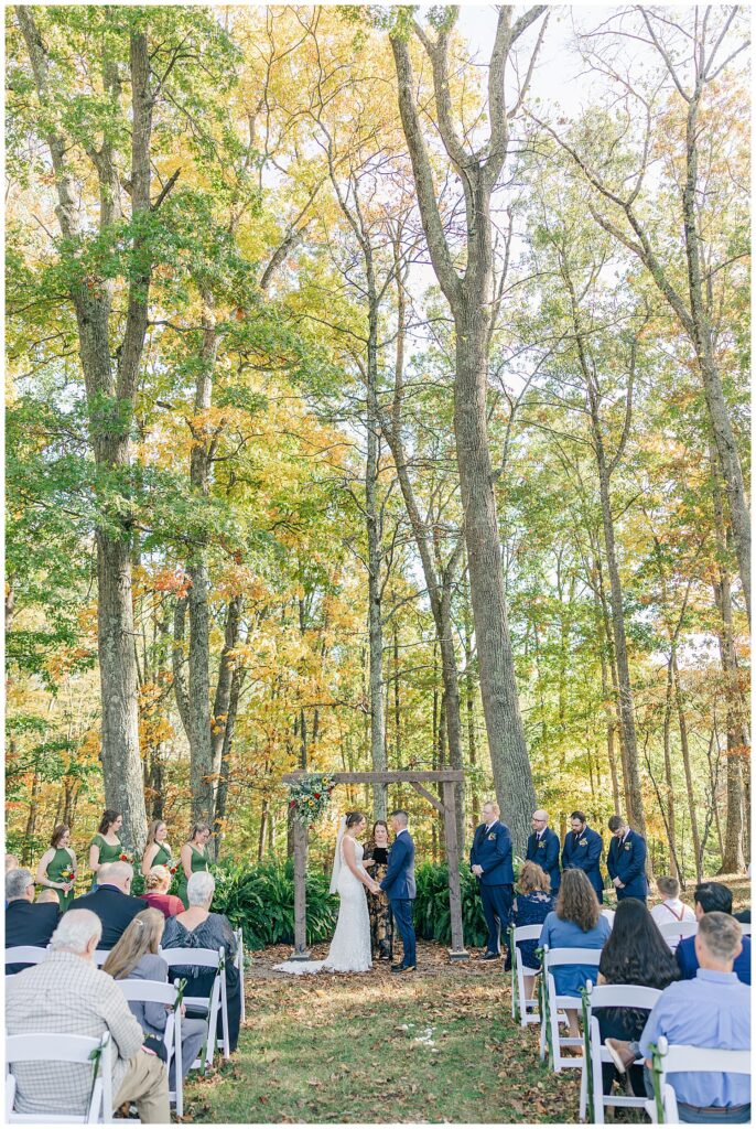 Guests seated outdoors watching the bride and groom exchange vows under a tall tree canopy with peak autumn color in the background.